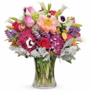 Colorful mixed‑flower arrangement featuring roses, peonies, hydrangeas, calla lilies, and seasonal blooms in a clear glass vase.